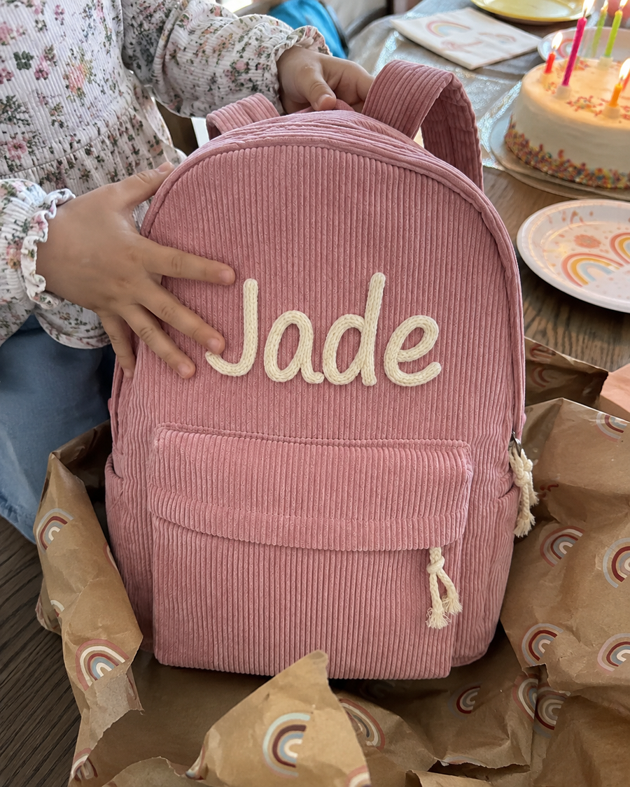 Sac à dos en velours côtelé rose avec le prénom Jade brodé main, déballé sur une table d'anniversaire, la main de l'enfant posée dessus