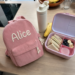 Photo client : sac à dos en velours côtelé rose avec le prénom Alice brodé main, sur la table de la cuisine avec la boîte à goûter