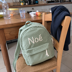 Photo client : sac à dos en velours côtelé vert sauge avec le prénom Noé brodé main, posé sur une chaise de cuisine au retour de l'école
