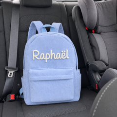 Photo client : sac à dos en velours côtelé bleu ciel avec le prénom Raphaël brodé main, posé sur la banquette arrière de la voiture