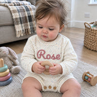Un bebe assis qui joue avec un anneau en bois, en barboteuse blanc mouchete au prenom Rose brode