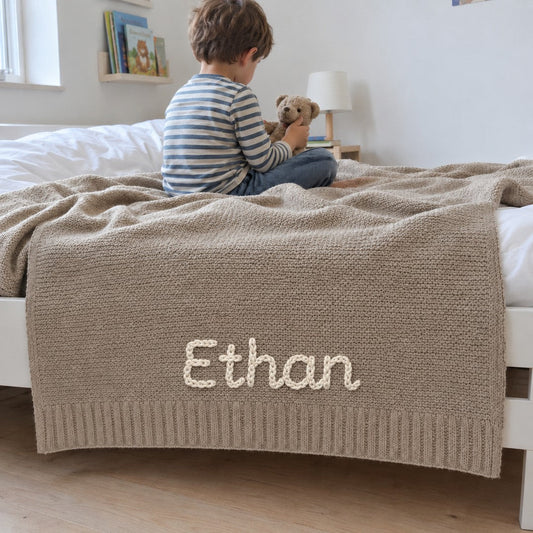Un enfant de quatre ans sur son lit, la couverture taupe brodee Ethan qui pend au bord du lit