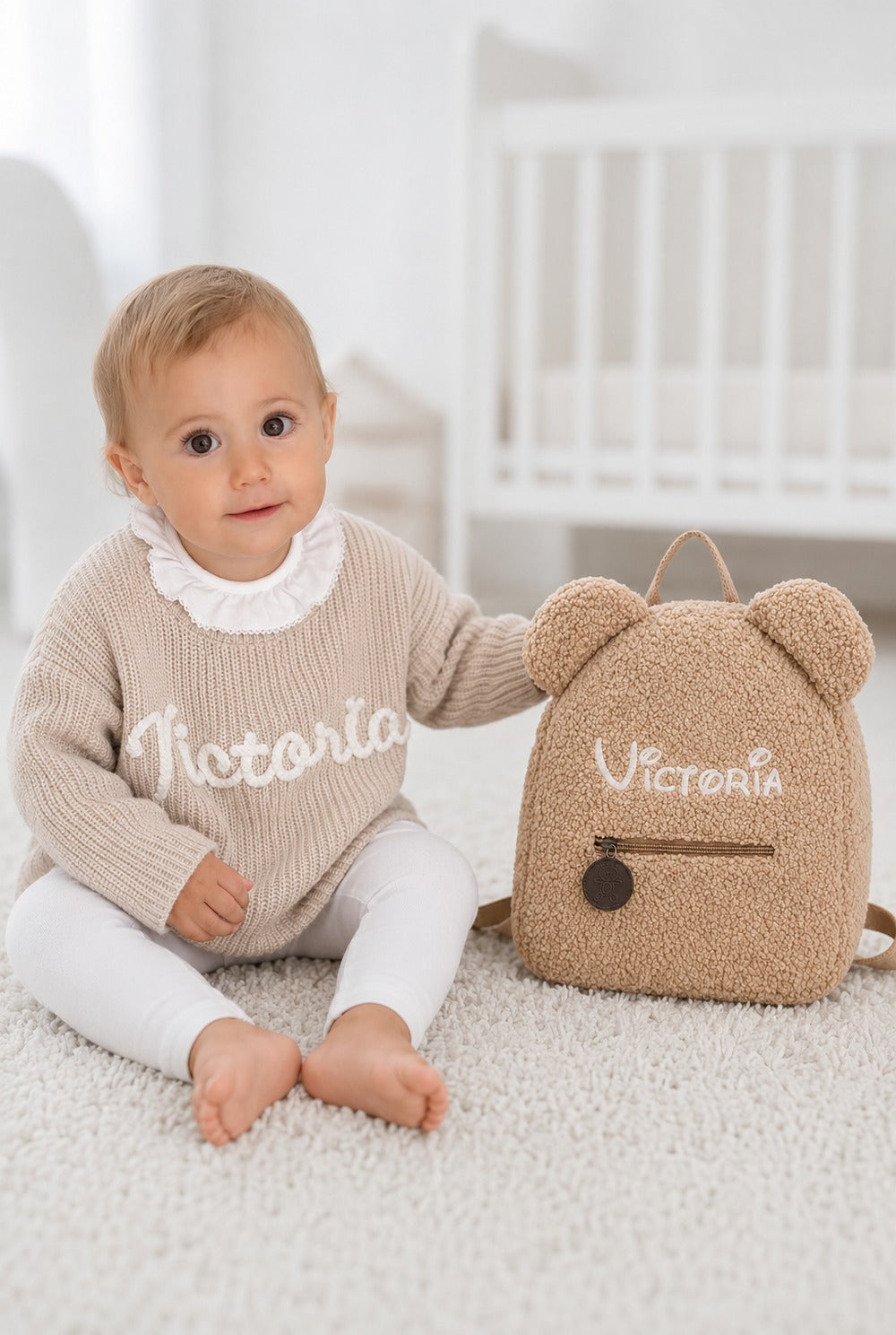 Petite fille en pull beige brodé à son prénom, assise à côté de son sac ourson personnalisé