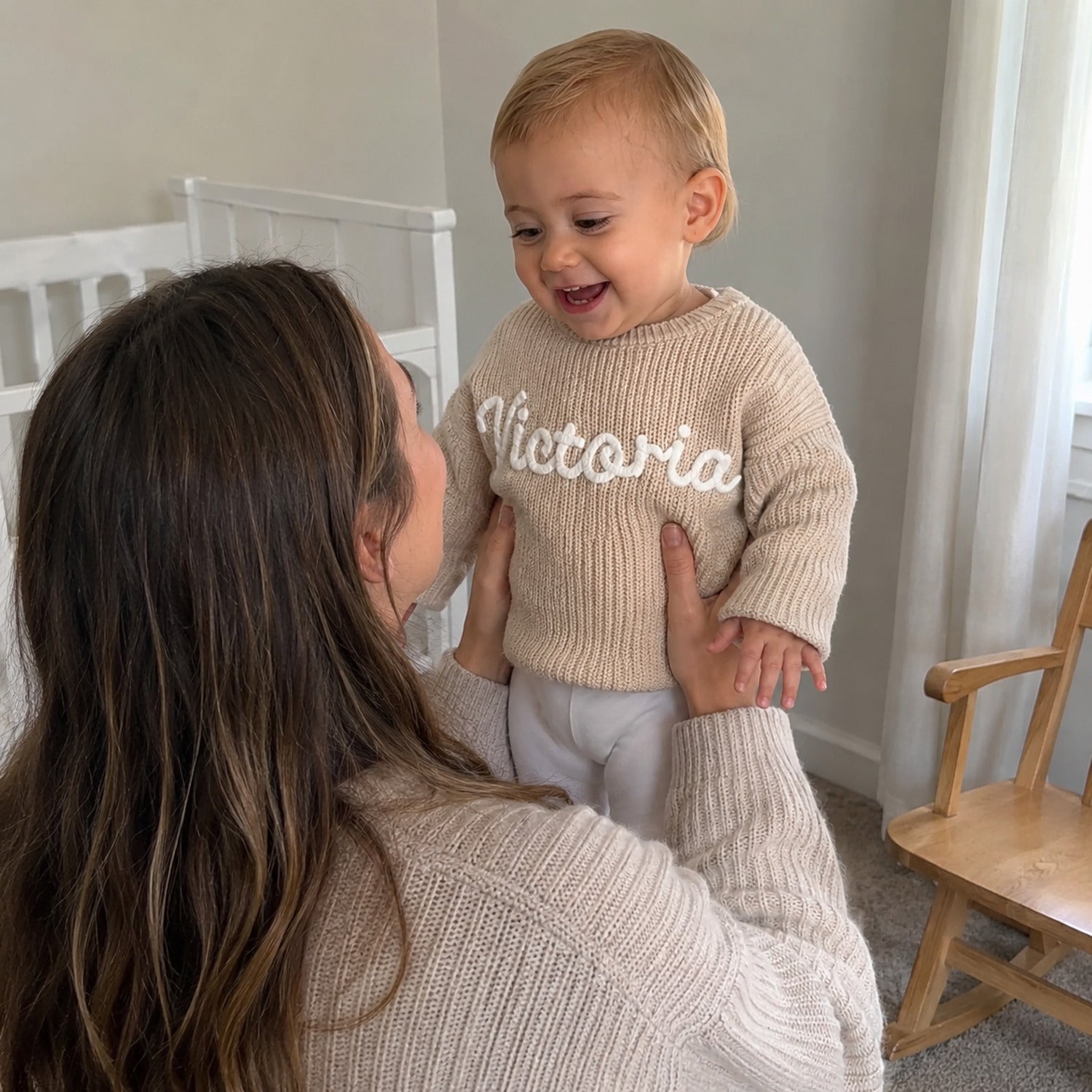 Maman qui souleve sa fille en pull beige brode a son prenom, dans la chambre