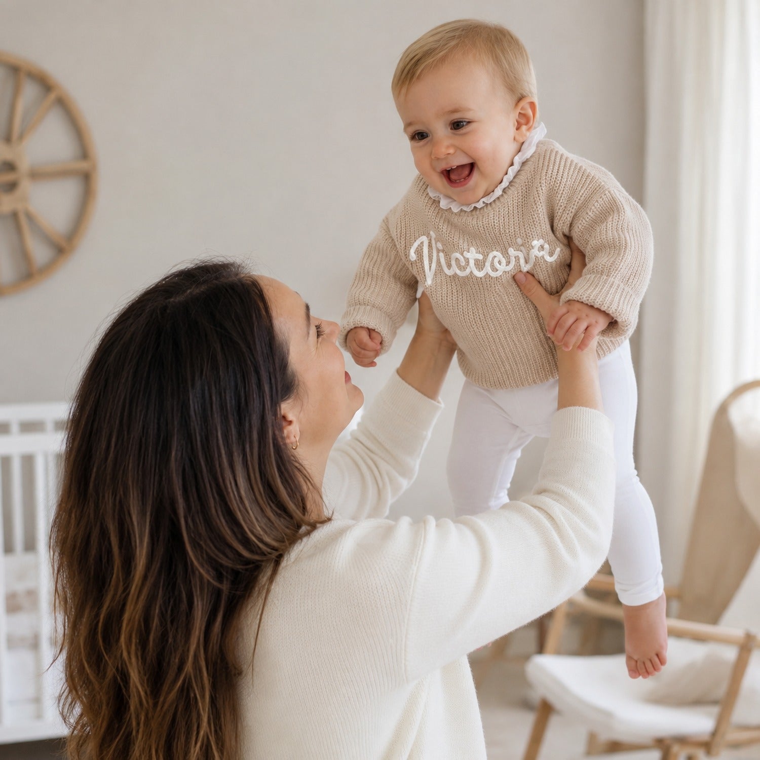 Maman qui soulève sa fille en pull beige brodé à son prénom, dans la chambre