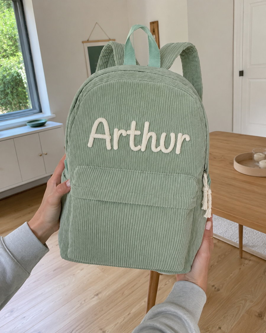 Sac à dos en velours côtelé vert sauge tenu à deux mains, prénom Arthur brodé main au point de chaînette en fil crème, salon clair