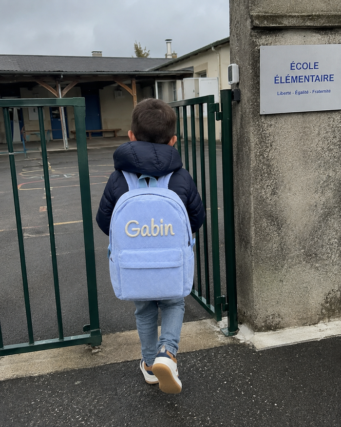 Garçon de dos franchissant le portail de l'école avec son sac à dos en velours côtelé bleu ciel, prénom Gabin brodé main en chaînette crème