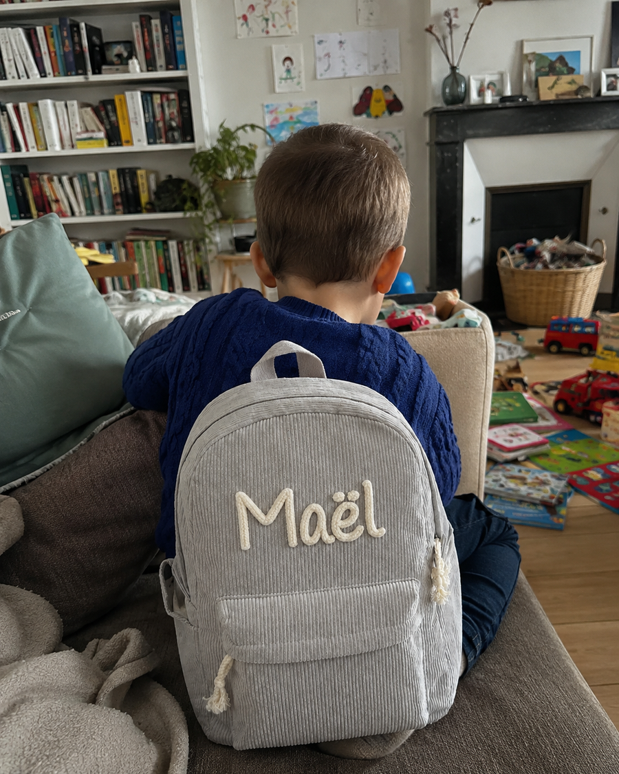 Garçon de dos assis dans le salon avec son sac à dos en velours côtelé gris, prénom Maël brodé main en chaînette crème, jouets et bibliothèque autour