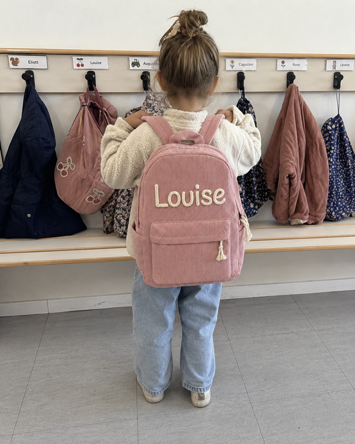 Petite fille de dos au vestiaire de l'école maternelle, sac à dos en velours côtelé rose sur les épaules, prénom Louise brodé main en chaînette crème, patères et sacs des autres enfants autour