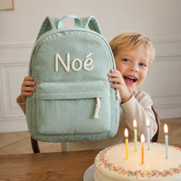 Garçon découvrant son sac à dos en velours côtelé vert sauge en cadeau d'anniversaire, prénom Noé brodé main en chaînette crème, gâteau et bougies devant lui