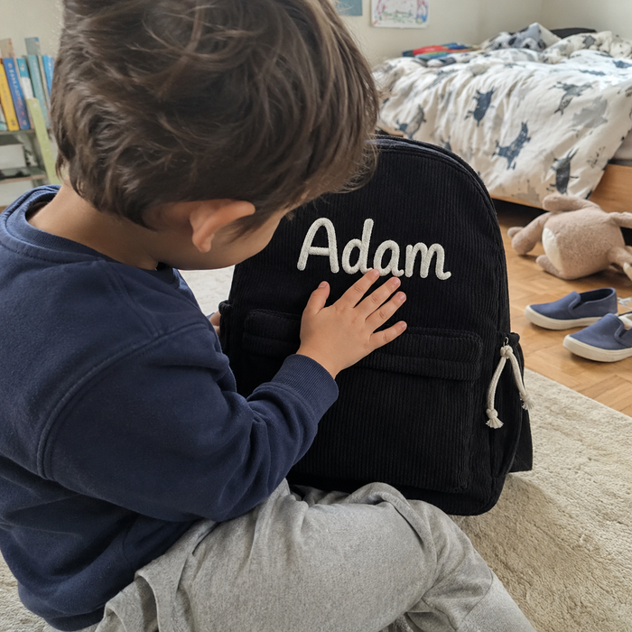 Garçon dans sa chambre, une main posée sur le prénom Adam brodé main en chaînette crème sur le sac à dos en velours côtelé noir