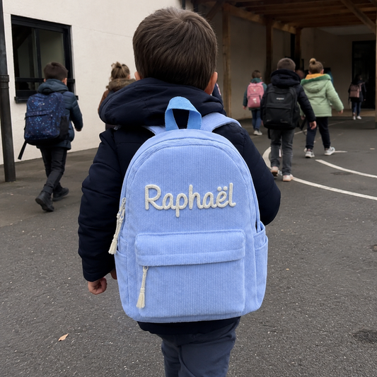 Garçon de dos entrant dans la cour de l'école maternelle avec son sac à dos en velours côtelé bleu ciel, prénom Raphaël brodé main en chaînette crème